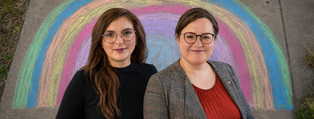 Zwei lächelnde Frauen stehen vor einem bunten Kreide-Regenbogen.